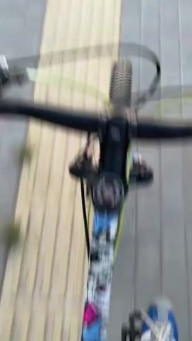 The video shows a first-person perspective of someone riding a bicycle along a paved path. The path is made of gray bricks with a textured yellow strip down the center. On either side of the path is a grassy area. In the distance, the sky is a pale orange and yellow, indicating either sunrise or sunset. There are some trees and utility poles visible. The footage is slightly blurry due to the movement of the bicycle.