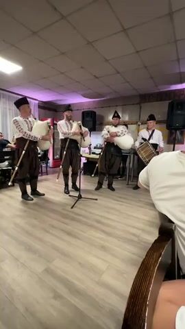 Four men in traditional outfits play musical instruments in a room with light wood floors and white walls. The three men on the left play bagpipes and the man on the right plays a drum. They are all wearing white shirts with embroidered patterns on the sleeves, dark vests, and dark pants. Two men on the left have black hats with a red band, and two men on the right have black hats. The men are standing in a semicircle with two people sitting at a table in the foreground. In the background, there are two large speakers and another person. The room is dimly lit with a purple light shining from the left.