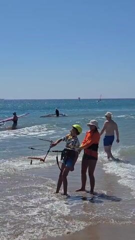 The video opens on a beach with two people standing in the shallow water, one holding onto a kite line. The person on the left is wearing a yellow helmet and a patterned shirt with blue shorts. The person on the right is wearing an orange top and dark bottoms. Behind them, other individuals are engaged in watersports. In the mid-ground, several people are on windsurfing boards, some holding masts and sails. In the background, the ocean stretches out to the horizon under a clear blue sky. The water is choppy with small waves breaking on the shore. The scene appears to be a sunny day at a beach popular for windsurfing and kiteboarding.