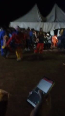 The video shows an outdoor performance at night. In the center, a person in a red jacket and yellow pants, wearing a headpiece of upright sticks, dances. A crowd of people surrounds the dancer, some standing and some sitting on orange chairs. In the background, a white tent is partially visible. In the foreground, a hand holds a lit-up smartphone. The ground appears to be dirt.