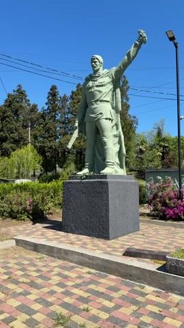The video shows a monument in a park-like setting.  A tall, light green statue of a man with his arm raised is on a dark gray, square base. The man is wearing a uniform and cape. The statue is in the foreground. Behind it is a green wall with "1941 1945" in red lettering. There is also a mural depicting soldiers. Several people are gathered near the wall, looking at something. The ground is paved with multi-colored bricks. There are bushes with pink flowers and trees in the background. The sky is blue.