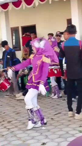A group of young people in marching band uniforms are preparing to perform. The lead performer, a young woman wearing a purple hijab and a purple uniform with gold trim, is in the foreground, facing left and extending her left arm. She wears white pants and tall purple boots with silver accents. Behind her, other performers are visible, some holding drums and wearing similar uniforms. The environment appears to be outdoors, with a paved ground and a building in the background. A white tent with red trim is set up overhead. Some people in the background are watching the performance. The overall scene suggests a lively event, possibly a parade or a school performance.