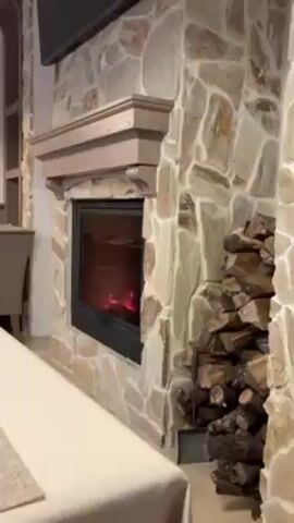 The video shows a close-up of a stone fireplace with a faux log fire inside. The fireplace has a wooden mantel and a black frame around the firebox. To the right of the fireplace is a stack of logs. The camera slowly zooms in on the faux fire.