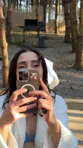A young woman is holding up her phone to take a selfie in an outdoor setting. She has long, dark hair and is wearing a white robe. A butterfly is perched on her forehead, near her eyebrow. The phone has a clear case with a circular magnetic charger visible, and a small sticker of a cat's face on the back. She is smiling and appears happy, perhaps amused by the butterfly.

The background suggests a wooded area, possibly a campsite or a rural backyard. Sunlight filters through the trees, indicating it is daytime. In the distance, there is a metal barbecue grill on a stand, and a gray trash can. The ground is covered in dry leaves and pine needles. The overall impression is a casual, pleasant moment in nature. The visible text on the phone screen is not legible.