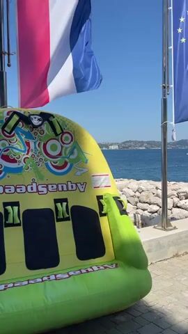 This media file captures a sunny day at Crveni Vrh, Croatia, likely during the summer. The scene is set by the Adriatic Sea, with a rocky shoreline and a clear blue sky. In the foreground, a bright yellow and green inflatable towable water toy, possibly for a "banana boat" ride, rests on the paved promenade. The toy has "Aquasense by" printed on it.

Behind the toy, two flagpoles stand tall. The first flagpole displays the French flag, recognizable by its blue, white, and red vertical stripes. Next to it, on the second flagpole, flies the blue flag of the European Union, adorned with its circle of yellow stars. Further down the promenade, a white Volkswagen Crafter van is parked, its license plate visible as "KR 855-MG".

The sea stretches out to the horizon, dotted with a few boats. The distant coastline features rolling hills covered in buildings, suggesting a town or resort area. The overall atmosphere is bright and cheerful, indicative of a pleasant, warm day by the sea, likely a tourist destination offering water sports. The presence of the French and EU flags could imply an international event or a location frequented by visitors from these regions.