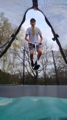 A person wearing a white shirt, shorts, black socks, and a yellow tie is seen indoors on a trampoline, from a low-angle view. The person is riding a pogo stick. The trampoline is enclosed by a net. Beyond the net are trees. The person jumps in the air on the pogo stick. The sky is visible above the trampoline.