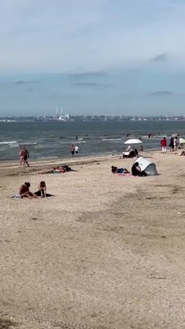 The video shows a busy beach on a sunny day.  People are sunbathing on the sand, some are lying on towels, and others are sitting.  There are several beach umbrellas and tents set up.  In the background, there are many people in the water swimming and wading.  The sea is calm with gentle waves.  In the distance, there is a cityscape with industrial buildings and ships visible on the horizon. The sky is partly cloudy.