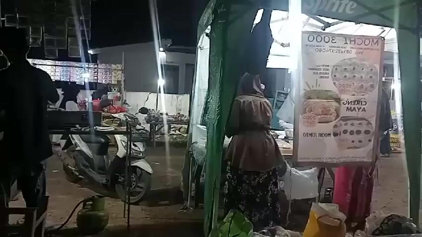 The video shows a night scene at an outdoor food stall. A woman wearing a brown top and a headscarf is standing behind a stall, reaching for something. To her right, a sign with Indonesian text, possibly advertising prices for food items like "MOCHI 3000", is visible. A motorcycle is parked to the left of the scene, with a green gas tank situated nearby. Further in the background, more stalls are illuminated by lights, and several people are present. The overall environment suggests a busy, informal marketplace or street food area.