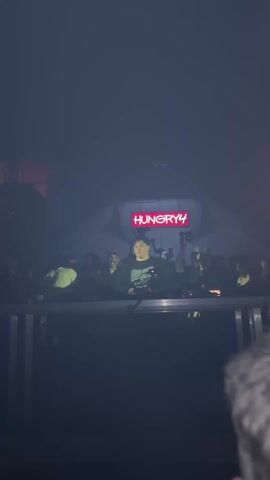 The video shows a DJ performing at a crowded event with the stage name “HUNGRY4” displayed in a red light sign. The DJ is standing behind a mixing console with other people around. The scene is dark and smoky, with several beams of light illuminating the space.  Some people are taking photos. The camera angle is from behind a person with short, light-colored hair.