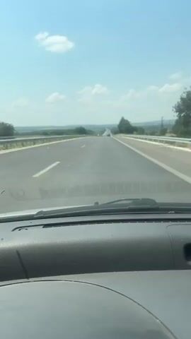 The video shows a view from inside a car driving on a highway. The road is asphalt and has lane markings. There are guardrails on the sides of the road. Trees and fields are visible in the background, along with a clear blue sky with some white clouds. Other vehicles, including a white van, can be seen on the highway ahead and to the left. The camera is positioned on the dashboard of the car.