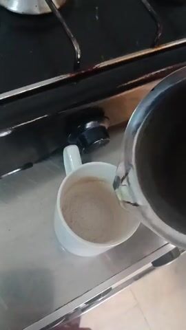A stainless steel pitcher is pouring a brown liquid into a white coffee mug on a stainless steel countertop. A gas stovetop is visible in the background.