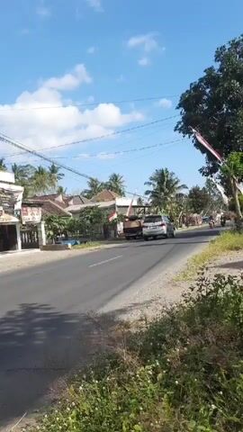 A daylight exterior shot shows a rural road with traffic. On the left, a row of buildings with white walls and tiled roofs lines the side of the road. Coconut trees are visible behind the buildings. A paved road runs horizontally across the frame, with a gravel shoulder on the right side covered in green vegetation. Several vehicles are on the road, including a truck, a car, and multiple motorcycles. One motorcycle with a rider passes by the camera from left to right. The sky is blue with scattered white clouds. Flags are visible attached to poles along the road.
