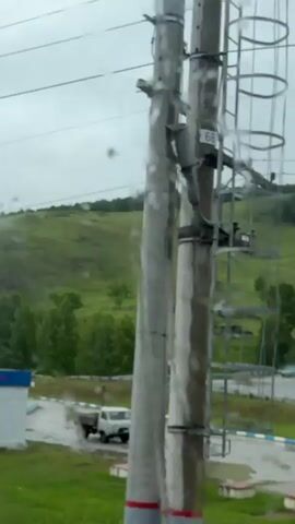 The video pans across a rural landscape under a cloudy sky. In the foreground, a white building with a blue roof is visible, along with a white truck. The middle ground features a lush green hill dotted with trees and small houses. The background consists of more rolling hills covered in greenery. The footage appears to be taken from a moving vehicle, possibly a train, as suggested by the presence of overhead wires and a slight blur from motion. There are some water droplets on the lens, giving the image a slightly diffused quality. No people are clearly visible in the frame.