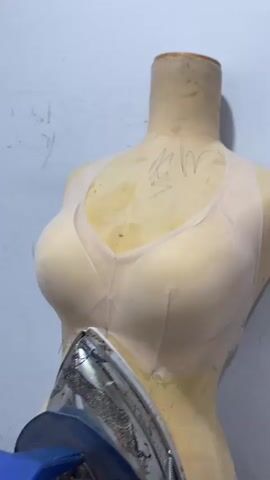 The video shows a close-up view of a dressmaker's mannequin on which a light-colored bra or bustier is being fitted. The mannequin is covered in fabric and has a faint drawing on its chest area. In the foreground, a hand holds a blue and silver steam iron, pressing it against the garment on the mannequin. The iron is positioned near the center of the chest area of the garment. The background appears to be a plain, light-colored wall. The focus is on the process of fitting and ironing the garment on the mannequin.