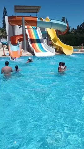 A sunny day at a water park. A large blue swimming pool is in the foreground, with several people splashing and swimming. In the background, a multi-colored water slide complex with orange, white, rainbow, and yellow slides is visible. The sky is clear and blue, and trees are visible behind the slides. There are people on the slides and in the pool, enjoying the water. One person is at the bottom of the rainbow slide and splashes into the pool. Another person is swimming towards the right side of the pool. Two people are playing in the water. A man is standing in the pool.