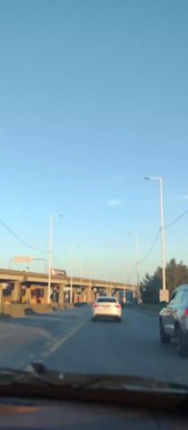 From inside a vehicle, a view down a paved road reveals two lanes marked by yellow lines. Ahead, two cars are visible, one white and one silver. Elevated above the road is a multilane highway or bridge, supported by numerous pillars and streetlights. A sign is affixed to the structure above. The sky is clear and blue, suggesting a sunny day.  Additional streetlights line the side of the road. A billboard promoting something as "DISTINTO" can be seen off to the right. The vehicle filming is in motion.