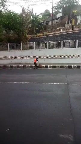 A person wearing an orange vest and a black hat is sweeping leaves with a broom on the side of a road. They are standing on a concrete surface next to a drainage ditch. Behind the person is a decorative wall with a metal railing on top. Further back, there are buildings and trees on a hillside. The road in the foreground is paved with asphalt and has white lines. The sky appears overcast.