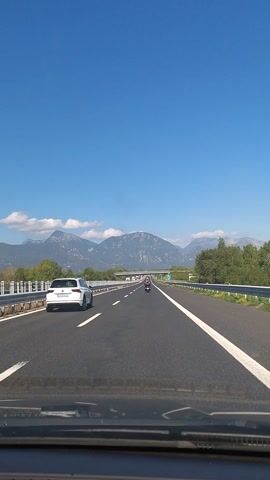 The video captures a view from inside a vehicle driving on a highway in Tomba, Italy. The scene is dominated by a clear, bright blue sky and a straight stretch of asphalt road. In the foreground, the dashboard of the vehicle is visible, suggesting the perspective of the driver or a passenger.

The highway has multiple lanes, indicated by white lane markings. Several vehicles are present, including a white car in the left lane and two motorcycles further ahead in the right lane. The road is lined with metal guardrails on both sides, separating the highway from the surrounding landscape.

Beyond the highway, the scenery features a backdrop of majestic mountains with soft, white clouds dotting the sky above them. Green trees and vegetation line the sides of the road, indicating a relatively rural or scenic area. The overall atmosphere appears peaceful and pleasant, suggesting good weather and daylight, likely mid-morning or afternoon given the bright sunshine and clear visibility.

A green highway sign with a white arrow indicating a turn or exit is visible on the right side of the road. The time and location context provided suggest this footage was recorded during daytime travel on an Italian highway. No explicit emotions are conveyed, but the clear skies and open road evoke a sense of journey and exploration. No readable text is discernible beyond the highway sign.