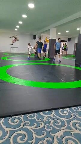 A group of young boys in athletic wear are practicing on a wrestling mat in a brightly lit room. The mat is black with several large green circles marked on it, indicating wrestling areas. The boys are moving around the mat, some walking, some appearing to be in motion as if practicing wrestling techniques. In the background, there are posters on the wall depicting wrestling figures and flags. The room appears to be a dedicated training facility with multiple wrestling mats visible. The flooring in the foreground is a patterned carpet.