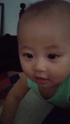 This is a close-up shot of a bald baby's head and shoulders. The baby is wearing a light green tank top with lace trim. The baby's eyes are open and looking downwards. The background is blurry and appears to be a room with a dark headboard of a bed visible.