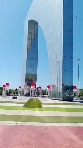 The video shows a shiny, metallic arch against a clear blue sky. The arch is made of individual, rectangular panels and has a large opening in the middle. At the base of the arch are several white, stylized figures with red hats. The area around the arch is a paved plaza with green patches of grass and landscaping. The camera pans upwards to capture the full height of the arch.