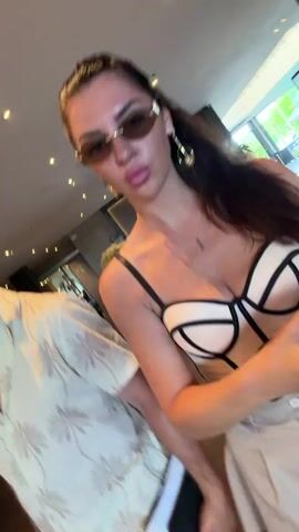 The video shows two people, a man and a woman, in what appears to be a hotel lobby. The woman is in the foreground, wearing a white and beige corset top and sunglasses. She is looking at the camera and smiling. The man is to her left, wearing a light-colored short-sleeved shirt with a floral pattern. He is also looking towards the camera. Luggage is visible in the background, and the lobby has a modern design with wooden floors and ceiling lights. The video appears to be a selfie taken by the woman.