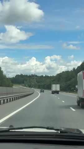 The video shows a view from inside a vehicle driving on a highway. The sky is blue with scattered white clouds. Trees line the right side of the highway, and a metal guardrail is visible on the left. Several vehicles are on the road, including a white truck in the right lane and two cars further ahead in the left lane. The road appears to be a multi-lane highway. The perspective suggests the camera is positioned behind the windshield.