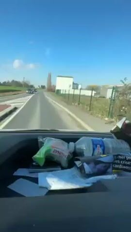 The video is taken from the dashboard of a vehicle moving along a paved road on a sunny day. The sky is a clear blue. Visible on the dashboard are a water bottle, a package of Daunat sandwiches, cigarette packaging, and some papers. Outside, the road is lined with grass and a white building with a white fence is visible on the right. Cars can be seen in the distance on the road ahead.
