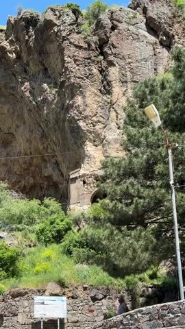 The video displays a mountainous landscape under a bright blue sky with scattered white clouds. Steep, rugged rock formations dominate the scene, with green vegetation, including bushes, trees, and patches of yellow flowers, growing on the slopes and at their base. A structure, possibly an ancient building or cave dwelling, is visible embedded within the face of one of the large cliffs. A winding road cuts through a green valley in the distance. In the foreground, a stone wall, a utility pole, and a coin-operated telescope are present. Several people are partially visible at the bottom of the frame, observing the scenery. The camera pans from left to right, showcasing the panoramic view of the natural environment.