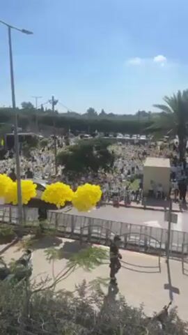 The video shows a large gathering of people dressed mostly in white, observed from an elevated position. The crowd is gathered in what appears to be a field or open area. Yellow balloons are tethered in the foreground, adding a pop of color. There are several parked cars, what appears to be a small stage, and other equipment on the outskirts of the crowd. A man dressed in dark clothing is running along a sidewalk. The weather is sunny and bright, and the sky is clear.