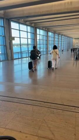 The video shows a wide, modern airport terminal hallway with a long glass wall on the right and large windows on the left, offering views of water and buildings. Sunlight streams in, indicating it's daytime, and the weather appears clear.

In the foreground, two individuals are walking away from the camera, each pulling a black suitcase. The person on the left is a man wearing a backpack, shorts, and a t-shirt. The person on the right is a woman in white wide-leg pants and a white top.

Further down the hallway, there is an escalator moving people upwards. Beyond the escalator, more people are visible walking and standing. A small white golf cart-like vehicle is parked on the left side of the hallway, with a person seated in it.

The overall atmosphere is that of a busy, yet orderly, transportation hub. The architecture is sleek and contemporary, with polished stone floors reflecting the light. The presence of luggage and the escalator suggests that people are traveling. No specific text is clearly legible in the video, other than a small green sign on the left with text that is too blurry to read. The location is identified as Tzafria, Israel.