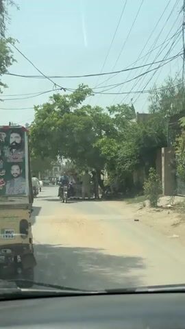 The video captures a street scene in Nasheman Colony, Pakistan, from the perspective of a moving vehicle. It is daytime, under a clear or slightly hazy sky, suggesting warm weather.

The main subjects are numerous men, predominantly dressed in traditional Pakistani attire (shalwar kameez), engaged in various activities. Several men are seen riding motorcycles on the paved but somewhat dusty road, while others are walking or standing.

On the left side of the frame, the front portion of a yellow auto-rickshaw or similar utility vehicle is visible, featuring a large advertising board with portraits of two men and some visible Urdu text, including the numbers "52144".

On the right side of the street, under the shade of a large green tree, there is a small vendor stall with red and white branding, where several men are gathered. Partially visible Urdu text on the stall appears to include "سپرائٹ" (Sprite), indicating it might be a beverage stand. Overhead, numerous power lines crisscross the sky. The overall scene depicts a typical bustling street environment in a residential or commercial area.
