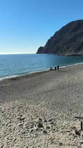 The video shows a beach with a view of the sea on a bright, sunny day.  The sand is a mix of light gray and darker patches, suggesting a combination of sand and pebbles. Several people are scattered along the beach, some walking, some standing near the water, and others appearing to be fishing, with fishing rods visible in the sand.  The sea is a clear blue color with gentle waves, and the sun reflects brightly on the water's surface.  A rocky cliff borders one side of the beach, and mountains are visible in the distance on the other side.  The sky is a clear, cloudless blue. The camera pans slowly across the scene.