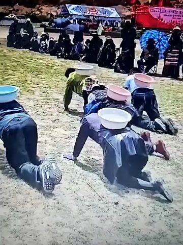 A group of people are on their hands and knees in a race, each balancing a white bowl on their head. Some people are wearing traditional clothing, while others are in modern attire. There are spectators in the background, some holding umbrellas. The ground appears to be dirt and grass. The overall atmosphere seems to be one of a community event or festival.