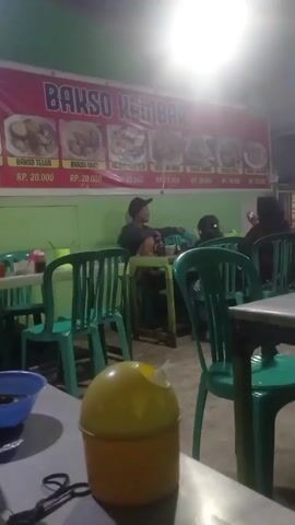 The video shows the interior of a restaurant with a banner in the background that reads "BAKSO GEMBAR." The banner displays pictures of various dishes with prices listed below them. Green plastic chairs are arranged around tables. Several people are seated at the tables, with some individuals facing away from the camera. One person is wearing a black cap. In the foreground, a yellow trash can is visible on a table. The lighting appears to be artificial and somewhat dim. The environment suggests a casual dining setting.