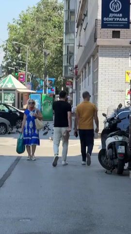A woman in a blue and white patterned dress walks on a sidewalk, carrying a green bag. Behind her, two men walk hand-in-hand away from the camera. The man on the left wears a black t-shirt and white pants, and the man on the right wears a yellow short-sleeved shirt and dark pants. Pigeons are scattered on the ground around them. In the background, there are buildings, trees, streetlights, traffic signals, cars, and a parked scooter. The scene appears to be a sunny day on a city street.