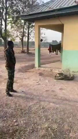The video shows a building exterior with a dirt yard. The building has a low-sloped metal roof and painted yellow and green walls. Through an open doorway, laundry can be seen hanging on a line. A man in camouflage uniform walks across the yard, pauses near the building, and then looks towards the field. The environment includes trees, and dry grass visible in the open area. The lighting suggests it is daytime.