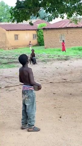 The video captures an outdoor scene in a rural village, likely in Riyom, Nigeria, focusing on a group of children engaged in various forms of play.

In the immediate foreground, a young boy, appearing to be about 8-10 years old, stands shirtless with his back slightly turned to the camera. He holds a light-colored, somewhat lumpy ball, possibly handmade, in his hands. He wears light blue, distressed jeans and dark sandals, with the word "NAYOVIN" faintly visible on his waistband.

To his right, another boy in a red t-shirt and light-colored pants stands observing the activity, hands clasped, wearing light green sandals. Further back in the mid-ground, two younger children are actively playing. One boy in a light brown shirt interacts with a black tire, while another child in a red shirt runs nearby. A fifth child, also in red, is visible closer to the houses.

The ground is unpaved, consisting of dry, light brown earth. The background features several simple, single-story houses with reddish-brown corrugated metal roofs and walls made of mud or brick. Sparse green grass grows around the houses, and lush tree foliage overhangs the top of the frame, providing shade.

The scene takes place during the daytime under bright but diffused light, suggesting mild weather. It conveys a sense of innocent childhood play and everyday life in a rural community.