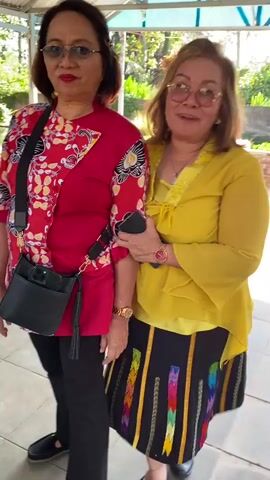 The video shows two women standing next to each other. The woman on the left is wearing a red and white floral shirt and black pants. She has a black purse slung across her body. The woman on the right is wearing a yellow top and a black skirt with colorful patterns. Both women are smiling and giving a thumbs-up gesture. The background appears to be an outdoor setting with some greenery and a roof overhead. The video is short and features a loop of the two women posing.
