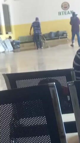 The video shows a waiting area with several people. In the foreground, there are several rows of black metal chairs with perforated seats. In the background, the floor is shiny and appears to be tile or polished concrete. Along the back wall, there is a yellow and white striped band, with the word "RTEAN" visible in green letters above a sign with a circular logo. Several people are visible. One person in a blue and white striped shirt is standing and leaning on a cane, facing away from the camera. Another person, also facing away from the camera and wearing a dark jacket and blue jeans, stands near a set of luggage. A third person, wearing a light blue patterned shirt and carrying a bag, walks away from the camera. Another person in a blue shirt is sitting on a bench. Various pieces of luggage are scattered around the waiting area. The lighting is bright, suggesting an indoor public space.