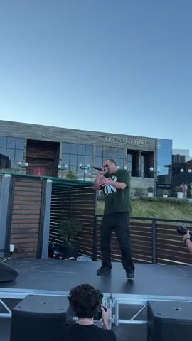 A man is performing on a stage outdoors, holding a microphone and wearing a green t-shirt with a white design and black pants. He appears to be a rapper or singer, with a confident stance and engaging in animated gestures. Several people are visible in the foreground, some holding cameras, suggesting they are filming or photographing the performance. In the background, a building with the text "LISI EVENT HALL" is clearly visible, indicating the location. The sky is clear and blue, suggesting it's daytime, likely late afternoon or early evening given the lighting. The overall atmosphere appears to be a lively event.