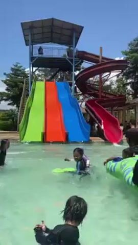 The video shows a water park on a sunny day. Several children in swimsuits and at least two adults, one of whom is wearing a black hijab, are standing in a shallow, light-blue pool, partially submerged to their waists. One adult holds a young child. Some children are sitting on a large, inflatable green and blue toy shaped like an animal. In the background, there are tall water slides with green, orange, blue and red sections, and an elevated platform structure. Near the edge of the pool, a gazebo and a few other people can be seen in the background, along with trees and other vegetation.  The overall scene depicts children having fun in a water park under clear skies.