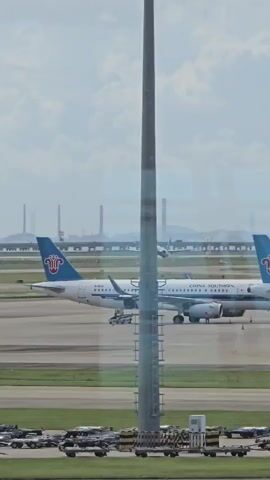 The video shows an aerial view of an airport tarmac on a cloudy day. A China Southern Airlines airplane is parked on the tarmac, with its tail visible. In the background, another airplane is taking off. In the foreground, a tall, grey pole stands vertically in front of the camera. The sky is hazy, and distant industrial structures can be seen on the horizon. Several rows of baggage carts are lined up on the tarmac in the foreground.
