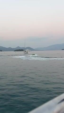 A watercraft with two people aboard is being towed across a body of water by a speed boat. The sky is a pale pink and orange, suggesting dawn or dusk. In the background, a white sailboat and a mountainous coastline are visible. The overall scene captures a recreational activity on the water.