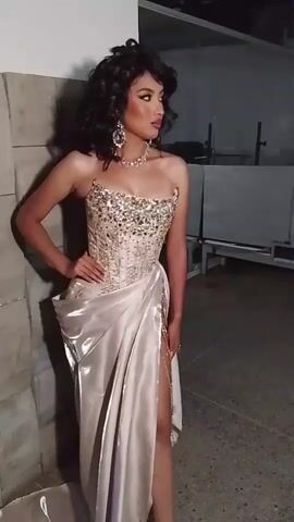 The video shows a person with dark curly hair, wearing a formal strapless light-colored dress with jewel embellishments on the bodice and a high slit, and ornate jewelry, standing in front of a wall that appears to be made of light gray blocks or tiles. They have one hand on their hip and are looking to their right. The background is somewhat industrial, with gray metal structures visible. The lighting is dim.