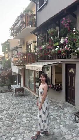 The video begins with a close-up of a young woman standing on a cobblestone street in front of a building adorned with balconies overflowing with flowers. She is wearing a white and blue patterned strapless dress and dark sandals. She smiles and gives a peace sign to the camera. The camera then pans to the left, revealing a wider view of the street, which is lined with shops and restaurants. Flags are visible on some of the buildings. In the background, there is a sunset casting a warm glow over the scene. The street is paved with large, irregular cobblestones. Further down the street, tables and chairs for outdoor dining are visible. The buildings have a mix of architectural styles, with some featuring balconies and awnings. The overall impression is a charming, historic street in a European town. The woman is the only person clearly visible in the foreground.
