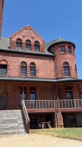 The video shows a large, old, red brick building with a tower and a turret. It is a sunny day with a clear blue sky. The building appears to be a historical landmark or an old institution. The camera pans across the facade, showcasing architectural details such as arched windows, stone accents, and a prominent staircase leading to an entrance. There are no people visible in the video. The environment appears to be outdoors, with some vegetation and a paved walkway in front of the building.