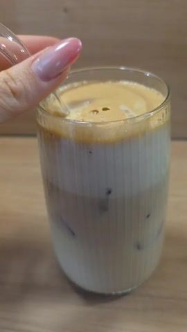 A hand stirs a glass of iced coffee on a wooden table. The glass is filled with ice, a light brown liquid, and a layer of creamy foam on top. The hand uses a clear straw to mix the ingredients, causing the foam to swirl and combine with the liquid below. The background is a plain wooden surface.