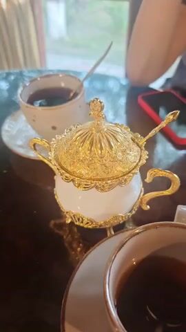 This media file shows a close-up of a white and gold sugar bowl, a cup of tea, and a person's hands on a dark, polished table. The sugar bowl is ornate, with detailed gold trim and a matching gold lid that rests beside it. A small gold spoon is inside the sugar bowl. Another cup of tea, with a spoon in it, is visible in the background. A red object, possibly a phone, is also on the table. The setting appears to be indoors, likely a cafe or restaurant, with a window and blurred background suggesting daylight. The lighting is soft, indicating it could be daytime. The scene conveys a sense of quiet enjoyment, possibly a tea break or a moment of relaxation. The focus is on the elegant sugar bowl and the act of preparing tea. No visible text is present. The context of Novy Urengoy, Russia, suggests a potentially cold climate outside, making the warm drink and cozy setting more inviting.