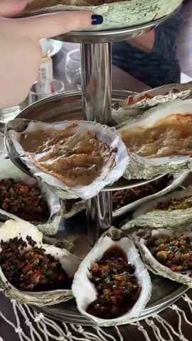 A tiered metal stand holds several oysters on a table. The oysters are in their shells, some appear to be raw, while others are cooked and topped with a relish or sauce. In the top left of the frame, a person's hand with a dark nail is reaching towards the top tier of oysters. The background is blurred but suggests an indoor setting, possibly a restaurant, with other tables and people visible. The lighting is warm and ambient.